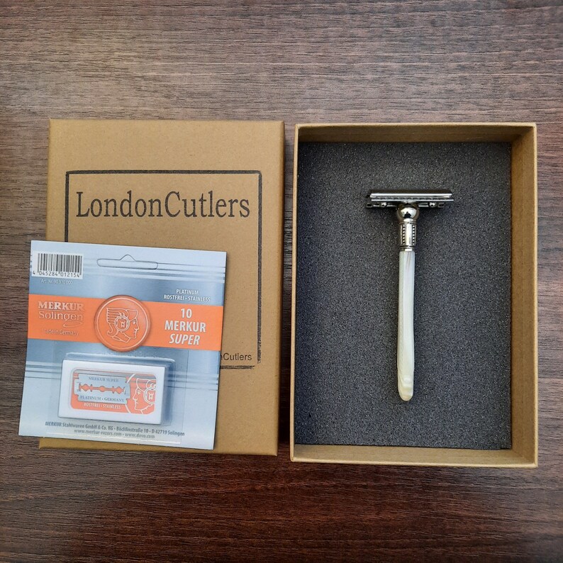 May include: A white and silver safety razor with a white handle in a brown cardboard box. The box has the words 'London Cutlers' printed on the front. The razor is packaged with a pack of 10 platinum coated razor blades. The blade packaging has the words 'Merkur Solingen' and '10 Merkur Super' printed on it.