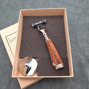 以下が含まれることがあります： 箱に入ったシェービングセット。セットには、茶色とシルバーのハンドルが付いたカミソリ、シルバーのスタンド、フォームインサート付きの箱が含まれています。カミソリのヘッドは黒とシルバーです。箱はベージュです。