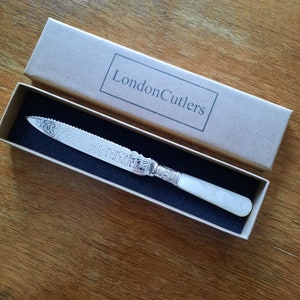 May include: A silver-toned serrated knife with an ornate floral engraved blade and a smooth white handle, presented in a rectangular cardboard box labeled LondonCutlers with a black foam insert. The knife features a decorative metal collar.