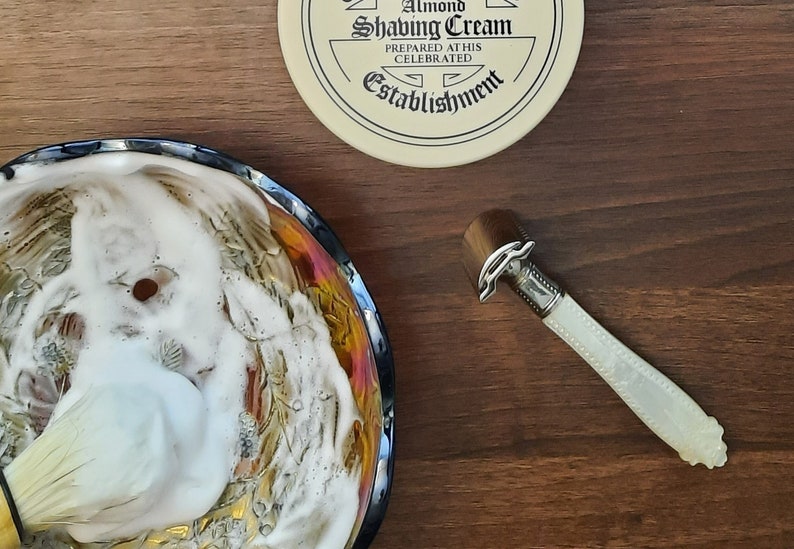 May include: A shaving brush with white lather in a decorative bowl, a safety razor with a white pearl handle, and a round container of shaving cream with the text 'Almond Shaving Cream Prepared at this Celebrated Establishment'.