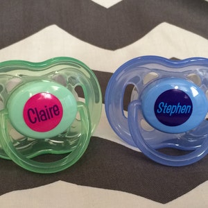 May include: Two pacifiers with personalized labels. One is green with a pink label that says "Claire" and the other is blue with a blue label that says "Stephen".