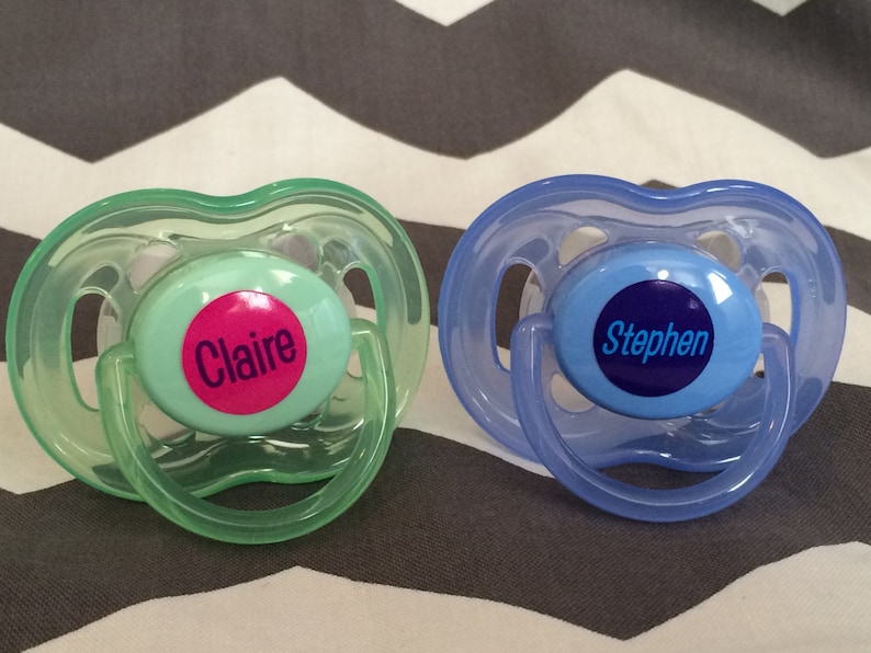May include: Two pacifiers with personalized labels. One is green with a pink label that says "Claire" and the other is blue with a blue label that says "Stephen".