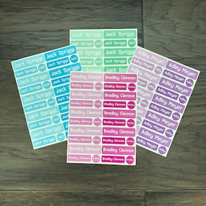 K&ouml;nnte beinhalten: Vier B&ouml;gen personalisierter Namensetiketten in verschiedenen Farben. Die Etiketten tragen Namen wie "Jack Spriggs", "Bradley Clennon" und "Adley Meyer". Die Etiketten sind rechteckig und oval, mit wei&szlig;er Schrift auf farbigem Hintergrund.