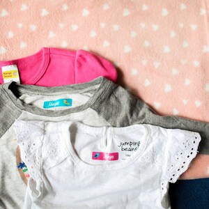 May include: Three shirts with colorful iron-on labels. The labels have the name "Maya" and a small bird design. One shirt is pink, one is gray with white sleeves, and one is white with lace trim.