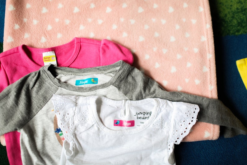 May include: Three shirts with colorful iron-on labels. The labels have the name "Maya" and a small bird design. One shirt is pink, one is gray with white sleeves, and one is white with lace trim.