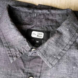 Puede incluir: Una camisa negra con cuello y una etiqueta blanca que dice "Layne Meyer" y "M" para el tama&ntilde;o.