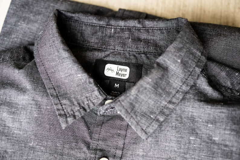 Puede incluir: Una camisa negra con cuello y una etiqueta blanca que dice "Layne Meyer" y "M" para el tama&ntilde;o.