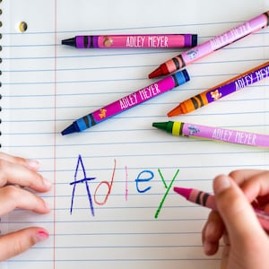 May include: A child's hand writing the name "Adley" in colorful crayon on a lined notebook. Several crayons with personalized labels reading "Adley Meyer" are scattered on the page.