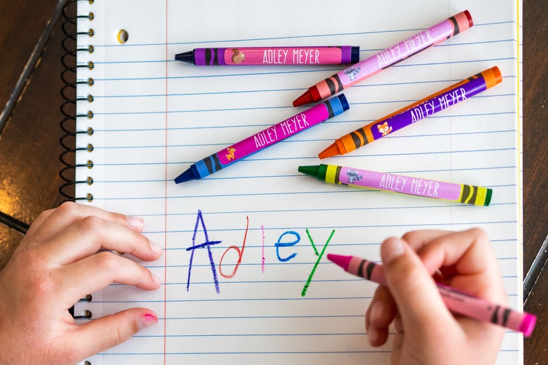 May include: A child's hand writing the name "Adley" in colorful crayon on a lined notebook. Several crayons with personalized labels reading "Adley Meyer" are scattered on the page.