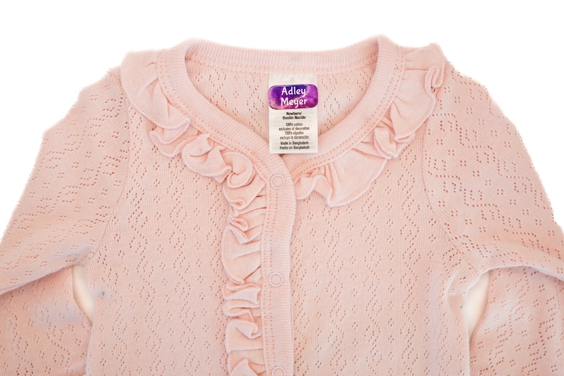 Puede incluir: Un c&aacute;rdigan rosa para beb&eacute; con cuello de volantes y cierre de botones. El c&aacute;rdigan est&aacute; hecho de un tejido de punto delicado y tiene una etiqueta que dice "Addley Meyer Boutique Knits".