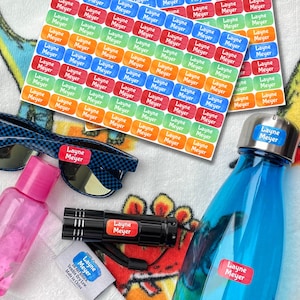 K&ouml;nnte beinhalten: Eine Auswahl personalisierter Etiketten mit dem Namen "Layne Meyer" in verschiedenen Farben, zusammen mit einer blau karierten Sonnenbrille, einer rosa Flasche, einer schwarzen Taschenlampe und einer blauen Wasserflasche. Die Etiketten sind rechteckig.
