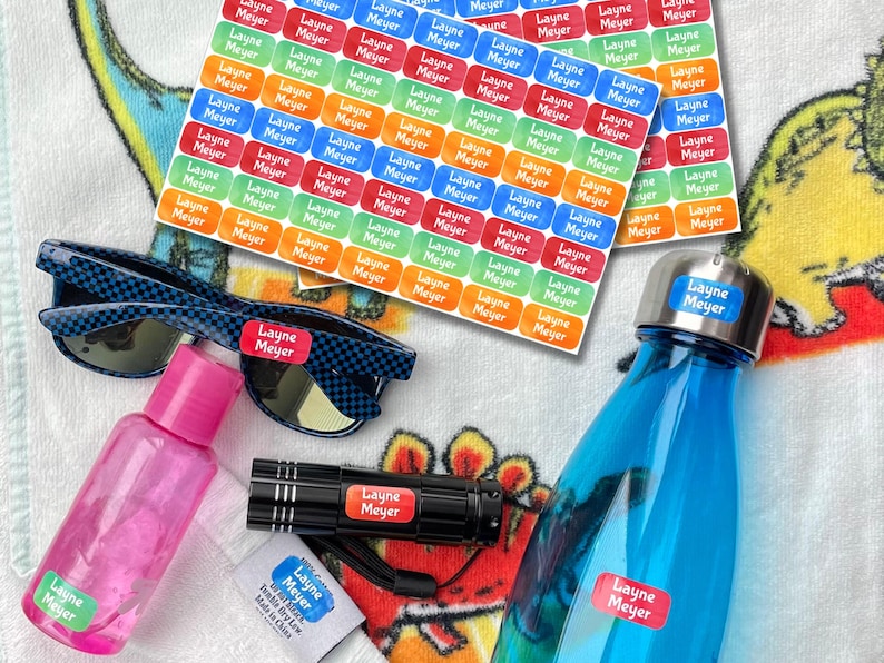 K&ouml;nnte beinhalten: Eine Auswahl personalisierter Etiketten mit dem Namen "Layne Meyer" in verschiedenen Farben, zusammen mit einer blau karierten Sonnenbrille, einer rosa Flasche, einer schwarzen Taschenlampe und einer blauen Wasserflasche. Die Etiketten sind rechteckig.