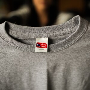 Puede incluir: Camiseta gris con una etiqueta roja y blanca que dice "Brody Matsu" y "Tweedway".