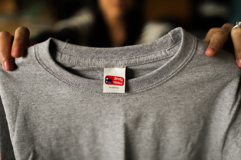 Puede incluir: Camiseta gris con una etiqueta roja y blanca que dice "Brody Matsu" y "Tweedway".