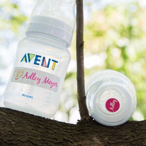 May include: Two clear baby bottles with labels. One bottle has a label that says "AVENT" and "Adley Meyer". The other bottle has a label that says "Adley".