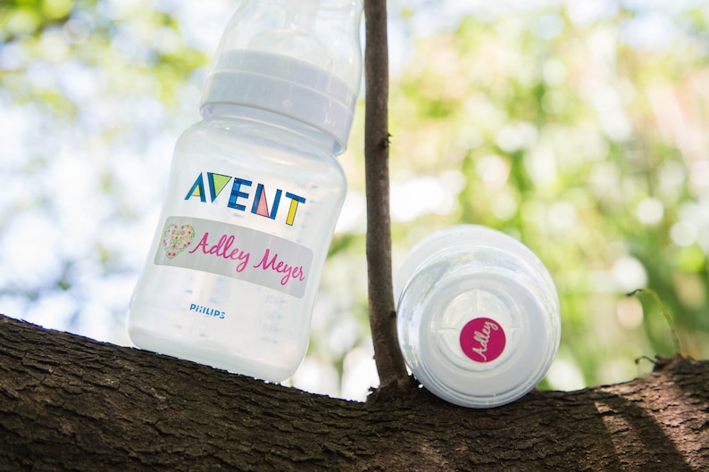 May include: Two clear baby bottles with labels. One bottle has a label that says "AVENT" and "Adley Meyer". The other bottle has a label that says "Adley".