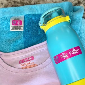 K&ouml;nnte beinhalten: Eine t&uuml;rkisblaue Wasserflasche mit gelbem Deckel und dem Namen "Allie Potter" auf einem rosa Etikett. Ein hellblaues Handtuch und ein rosa T-Shirt mit dem Namen "Allie Potter" sind ebenfalls im Bild.