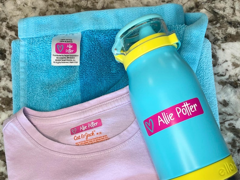K&ouml;nnte beinhalten: Eine t&uuml;rkisblaue Wasserflasche mit gelbem Deckel und dem Namen "Allie Potter" auf einem rosa Etikett. Ein hellblaues Handtuch und ein rosa T-Shirt mit dem Namen "Allie Potter" sind ebenfalls im Bild.