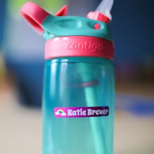 May include: A turquoise blue water bottle with a pink lid and a clear straw. The bottle has a pink label that says "contigo" and a purple label with a rainbow and the name "Katie Brewer".
