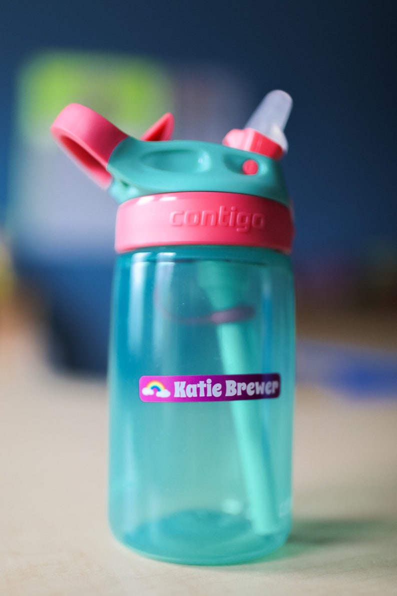 May include: A turquoise blue water bottle with a pink lid and a clear straw. The bottle has a pink label that says "contigo" and a purple label with a rainbow and the name "Katie Brewer".