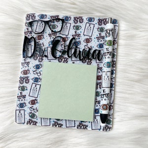May include: A white notepad with a black eye chart pattern and a black pen with a white eye chart pattern. The notepad has a green sticky note and a black acrylic nameplate that says "Dr. Olivia".