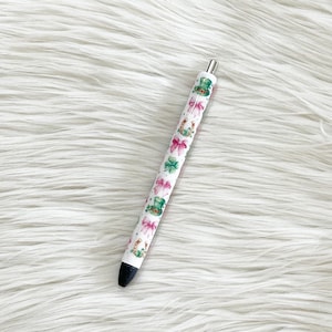 May include: A white pen with a black tip and silver accents. The pen is decorated with a pattern of pink bows, green shamrocks, and green leprechaun hats. The pen is laying on a white, fluffy surface.