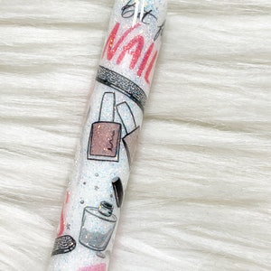 Peut inclure: Un stylo blanc avec un capuchon argent&eacute; et un design paillet&eacute;. Le stylo a un design rose et noir avec les mots "Get to the Nail" imprim&eacute;s dessus.