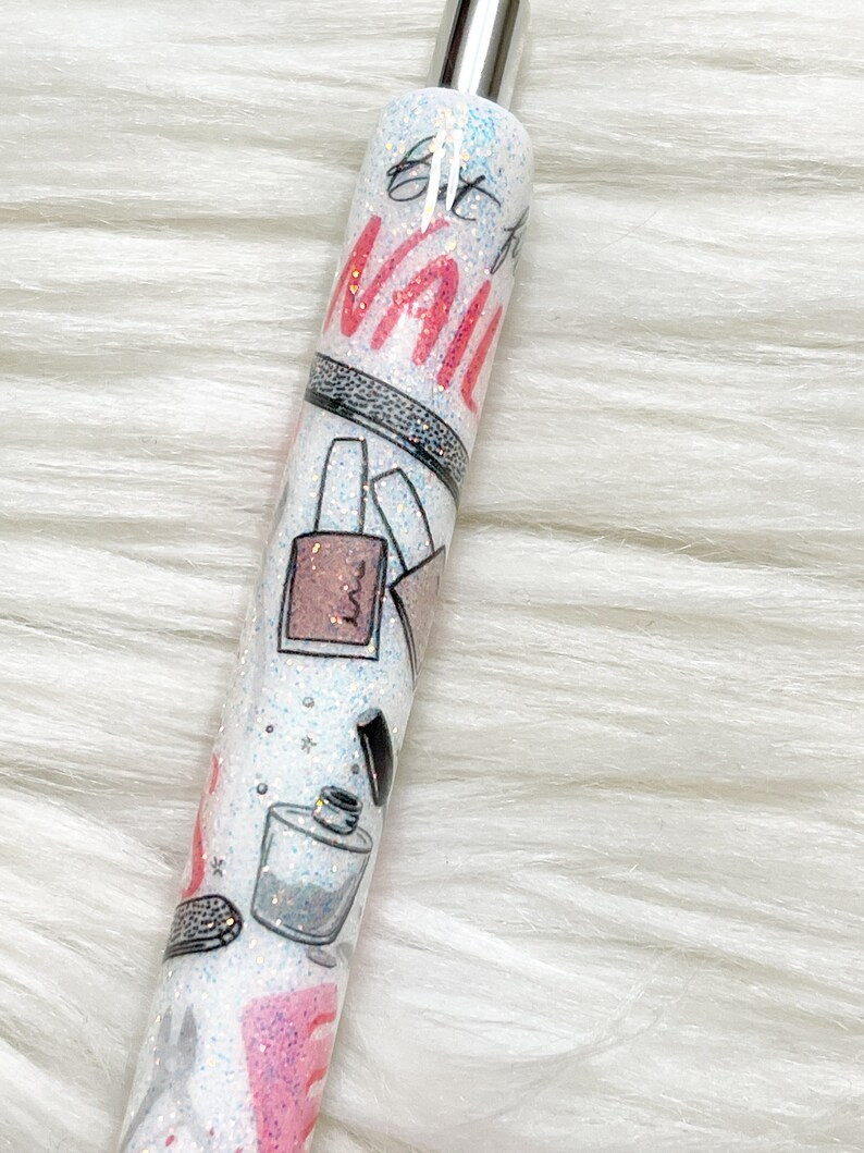 Peut inclure: Un stylo blanc avec un capuchon argent&eacute; et un design paillet&eacute;. Le stylo a un design rose et noir avec les mots "Get to the Nail" imprim&eacute;s dessus.
