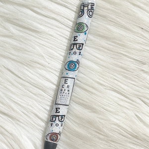 May include: A white pen with a glittery finish, decorated with eye charts, eyeglasses, and eye illustrations. The pen has a black tip and silver accents. The design includes the letters E, F, P, T, O, Z, and other letters from the eye chart.