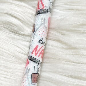 Peut inclure: Un stylo blanc avec une finition paillet&eacute;e et un motif rose et noir repr&eacute;sentant des flacons de vernis &agrave; ongles, des limes &agrave; ongles et le texte "But First, Nails".