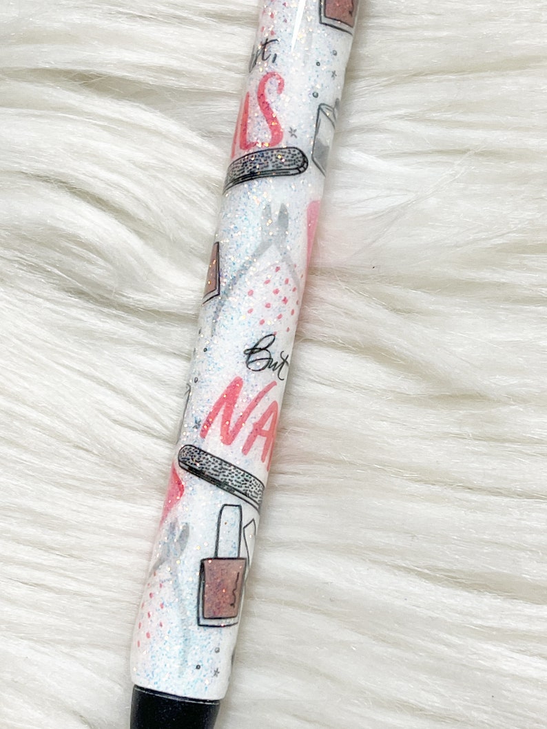 Peut inclure: Un stylo blanc avec une finition paillet&eacute;e et un motif rose et noir repr&eacute;sentant des flacons de vernis &agrave; ongles, des limes &agrave; ongles et le texte "But First, Nails".