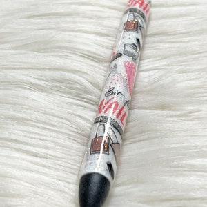 Peut inclure: Un stylo noir et blanc avec une finition scintillante et un motif rose et blanc. Le stylo porte l'inscription "But First, Coffee".