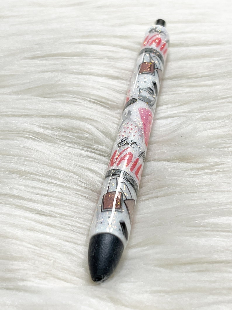 Peut inclure: Un stylo noir et blanc avec une finition scintillante et un motif rose et blanc. Le stylo porte l'inscription "But First, Coffee".