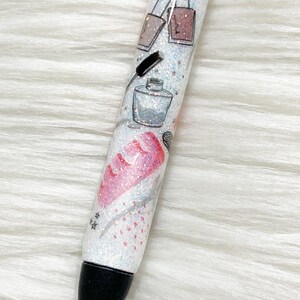 Peut inclure: Un stylo blanc avec une pointe noire et des paillettes incrust&eacute;es dans la peinture blanche. Le stylo a un motif de flacons de vernis &agrave; ongles, un pinceau &agrave; maquillage et un flacon de parfum.