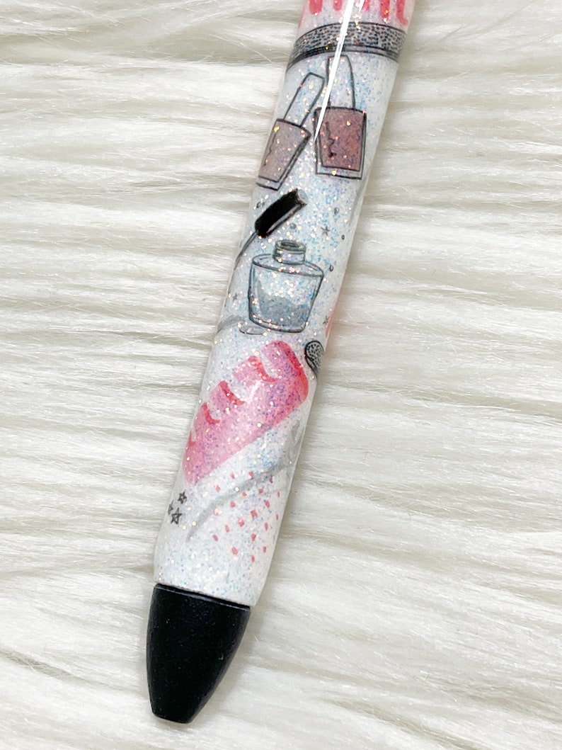 Peut inclure: Un stylo blanc avec une pointe noire et des paillettes incrust&eacute;es dans la peinture blanche. Le stylo a un motif de flacons de vernis &agrave; ongles, un pinceau &agrave; maquillage et un flacon de parfum.
