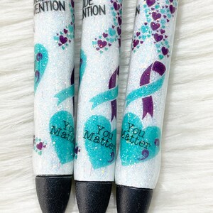 以下が含まれることがあります： ターコイズとパープルのリボンデザインとグリッターが施された白いペンが3本。ペンには「自殺予防」と「あなたは大切です」の文字が印刷されています。