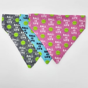 May include: Three dog bandanas with a tennis ball pattern. The bandanas are in pink, blue, and gray. The text on the bandanas reads "Ball is Life".