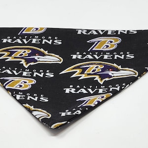 May include: Black bandana featuring the Baltimore Ravens logo and team name in white and gold. The bandana is triangular in shape and made of fabric. The Ravens logo is a purple raven head.