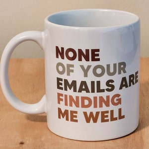 May include: White ceramic coffee mug with the text "NONE OF YOUR EMAILS ARE FINDING ME WELL" in brown, gray, and orange lettering.