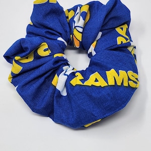 May include: A blue scrunchie with a repeating pattern of yellow and white text that reads "RAMS". The fabric is gathered and knotted to form a circular hair accessory. The scrunchie is on a white background.