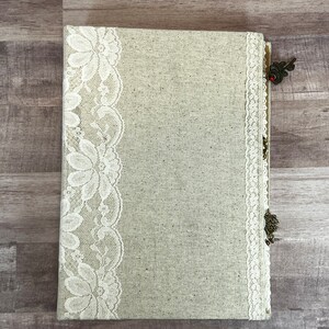 May include: A beige linen tablecloth with white lace trim and a brown tassel. The tablecloth is folded in half and laid flat on a wooden surface.