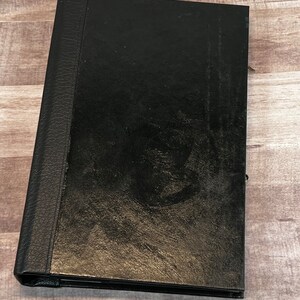 May include: A black leather-bound photo album with a textured spine and a slightly worn surface. The album is closed and lies on a wooden surface, suggesting a vintage or used item. The album's design is simple and classic.