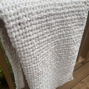 May include: A white crocheted blanket with a textured, fluffy surface. The blanket is draped over a wooden railing.