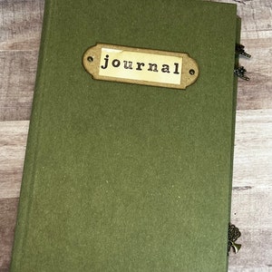 May include: A green journal with a brown label that says "journal" on it. The journal has a decorative metal charm attached to the spine.