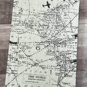 May include: A rectangular, off-white canvas-covered journal featuring a vintage world map design in black. The map includes continents, oceans, and flight paths, with the text "THE WORLD" and "MERCATOR PROJECTION". The journal has a classic, travel-themed aesthetic.