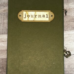 May include: A dark green journal with a brown label that says "journal" on it. The journal has a metal clasp and a decorative charm attached to it.