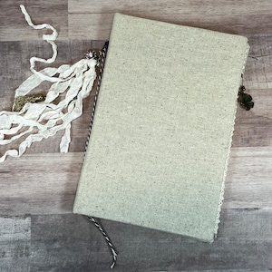 May include: A beige linen journal with a lace trim and a decorative tassel. The journal has a black and white striped ribbon bookmark.