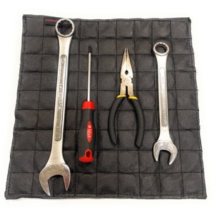 May include: A black leather tool mat with a grid pattern. The mat has four tools on it: a pair of pliers, a screwdriver, and two wrenches. The wrenches are silver and have black handles. The screwdriver is red and black. The pliers are silver with black handles.