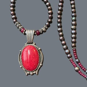 May include: A silver necklace with a red gemstone pendant. The necklace is made of small silver beads and has a long, thin chain.