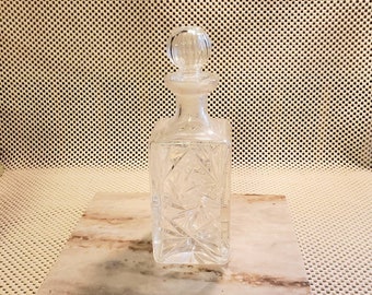 May include: A clear glass decanter with a square body and a round stopper. The decanter has a cut glass design and is sitting on a marble surface.